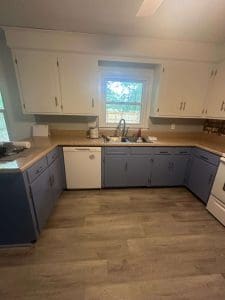 Freshly painted kitchen with white upper cabinets, blue-gray lower cabinets, window above sink, and wood-look flooring