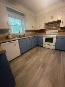 Freshly painted kitchen with white upper cabinets, blue-gray lower cabinets, brick backsplash, and wood-look flooring