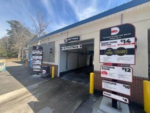 Freshly painted exterior of The Wash Zone Car Wash with Soft Touch bay, pricing signs, and tan stucco walls with red brick base